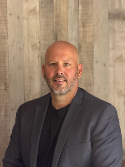 A bald man with a beard wearing a dark blazer and black shirt against a wooden background.