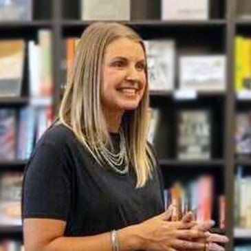 Smiling woman with layered necklaces in front of bookshelf.