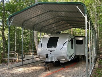 Traveler trailer parked under a metal carport in a wooded area.
