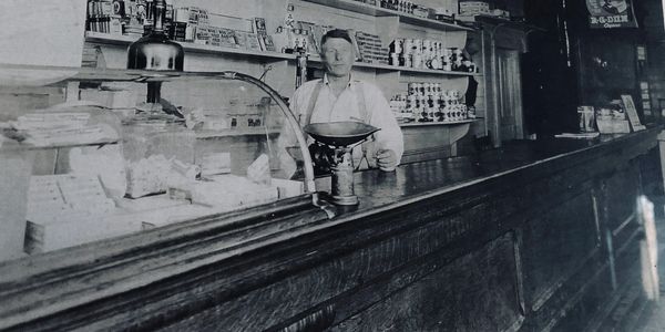 Black and white photo of a shopkeeper behind a wooden counter with products on shelves.