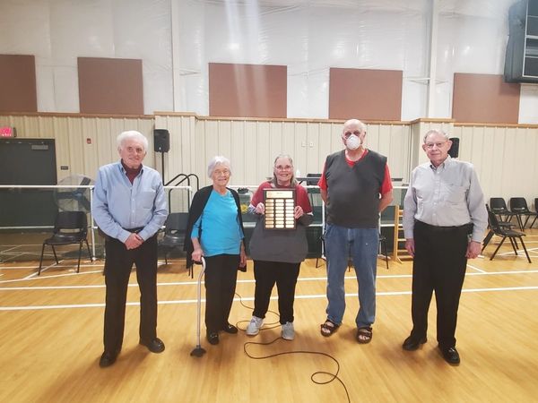 Volunteers of the Year for all past years and 2021 stand in a line in Rec Room.  Pam  H holds plaque