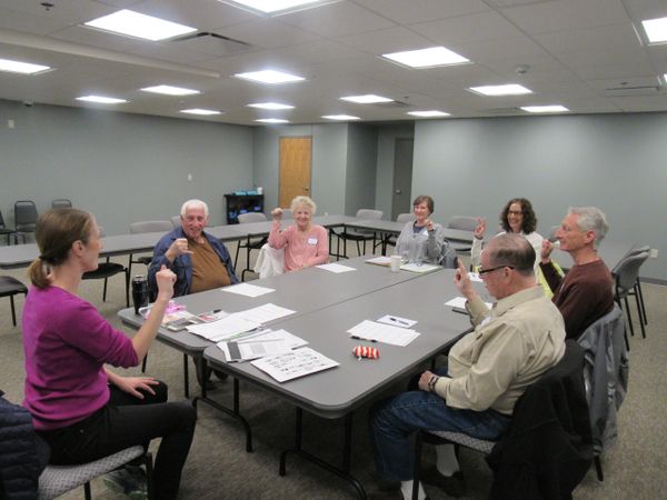 Sign Language instructor leads group around table learning to sign first letter of name