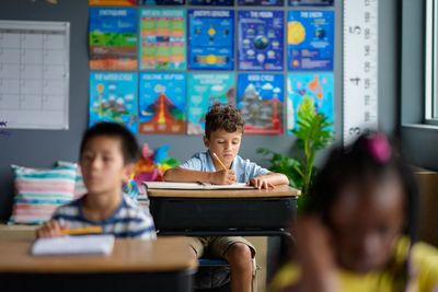 A young boy focused on writing in a classroom with colorful educational posters behind him.