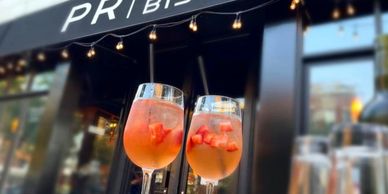 Two glasses of pink cocktails with fruit garnishes outside a bistro.