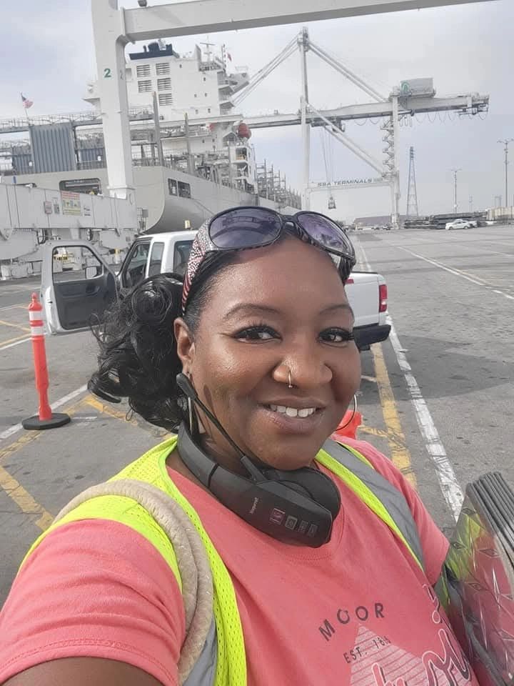 Smiling woman in a safety vest at a busy port with cargo cranes in the background.