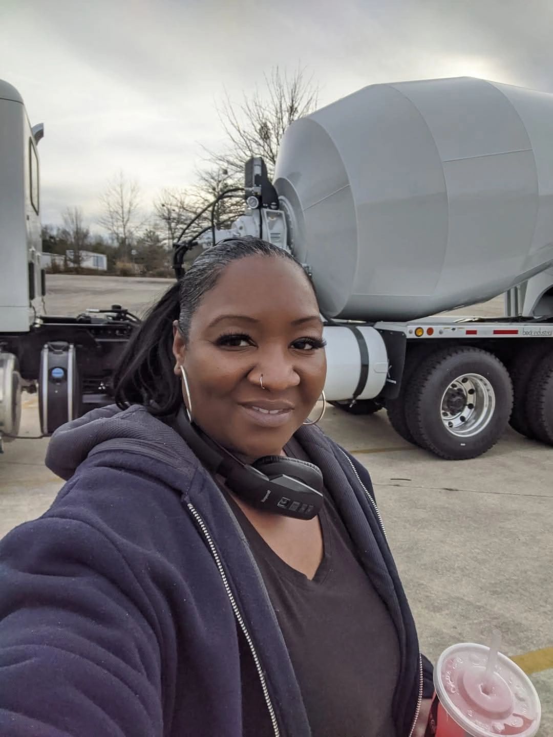 Woman taking a selfie in front of a cement mixer truck holding a drink.