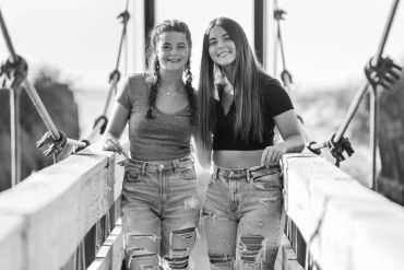 Two smiling young women standing on a wooden bridge.
