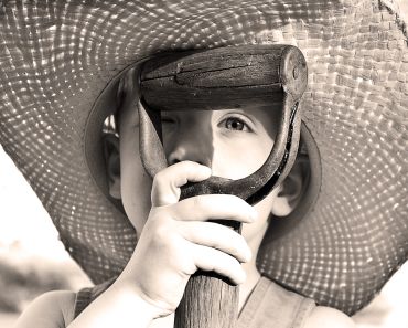 Child holding a wooden tool handle, wearing a large hat.