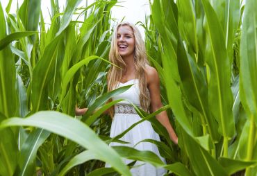 Smiling woman in white dress among tall green corn plants.