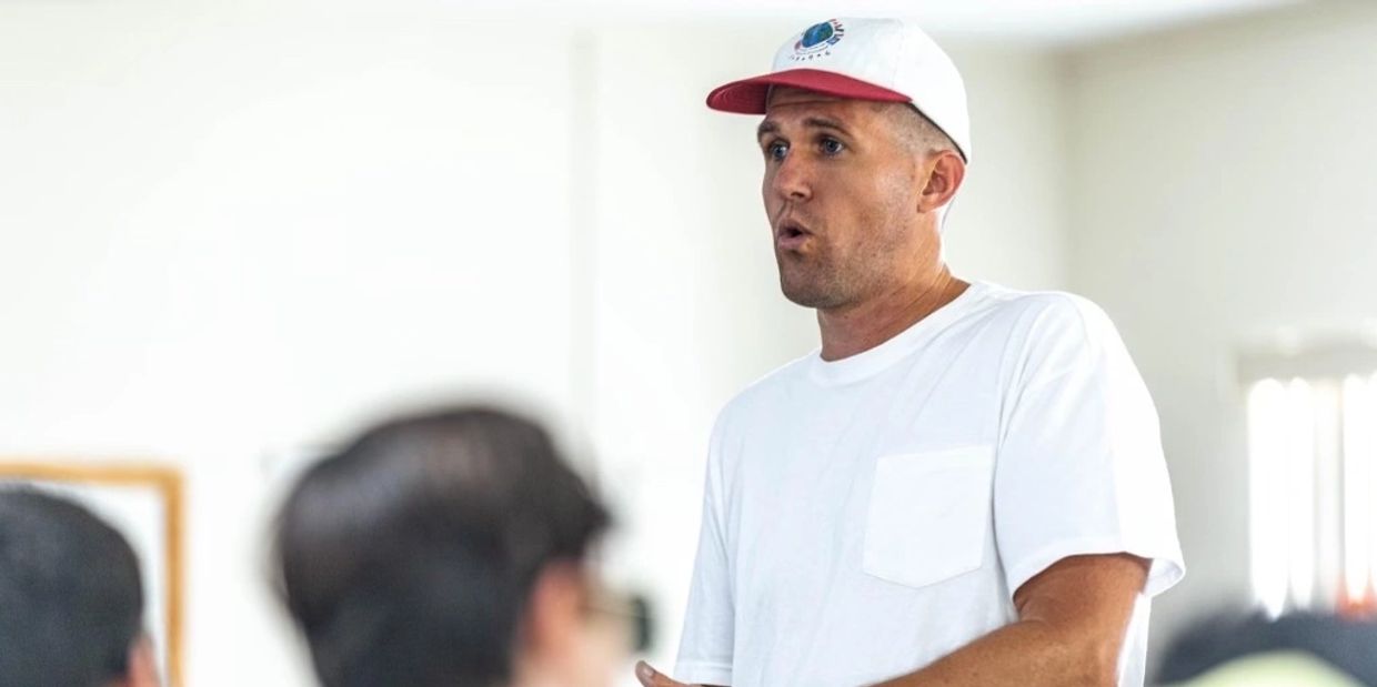 Man in white shirt and cap speaking to a group indoors.