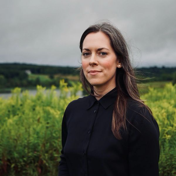 Woman in black shirt standing outdoors with greenery in the background.