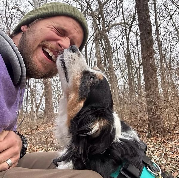 A man laughing as a dog affectionately licks his face outdoors.