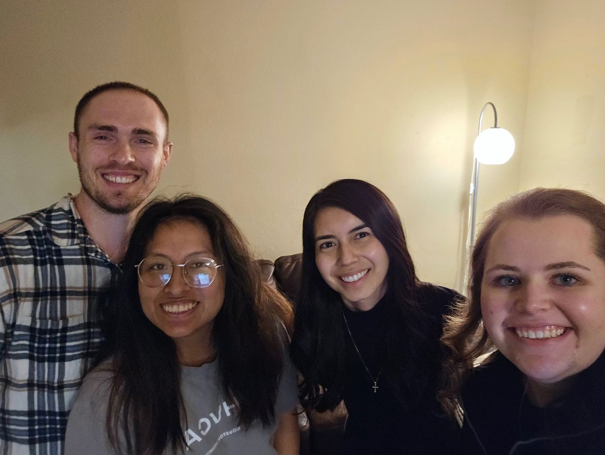 Four friends smiling indoors with a lamp in the background.