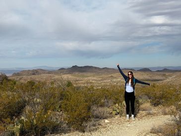 Woman joyfully posing with arms raised in a desert landscape under a cloudy sky.