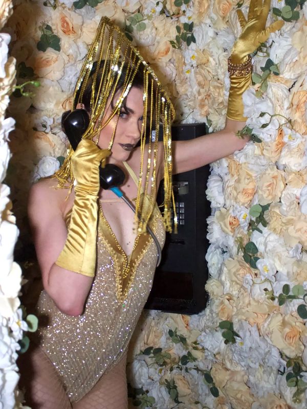 Person in sparkling gold outfit and gloves using a vintage phone surrounded by white and peach roses.