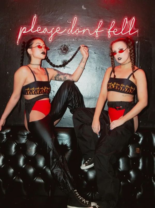 Two women in matching flame tops and red sunglasses pose under neon sign.