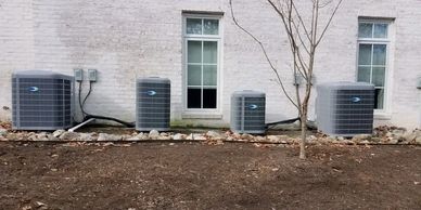 Four air conditioning units installed outside a white brick building with two windows and a tree.