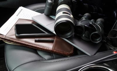 A camera, binoculars, laptop, phone, and documents on a leather car seat.