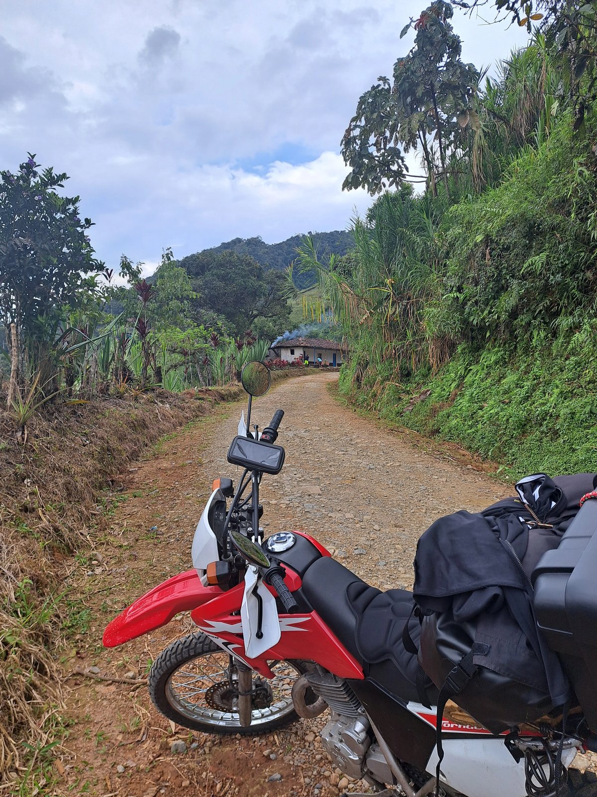 Motorcycle parked on a rural dirt road with lush greenery and a small house in the background.