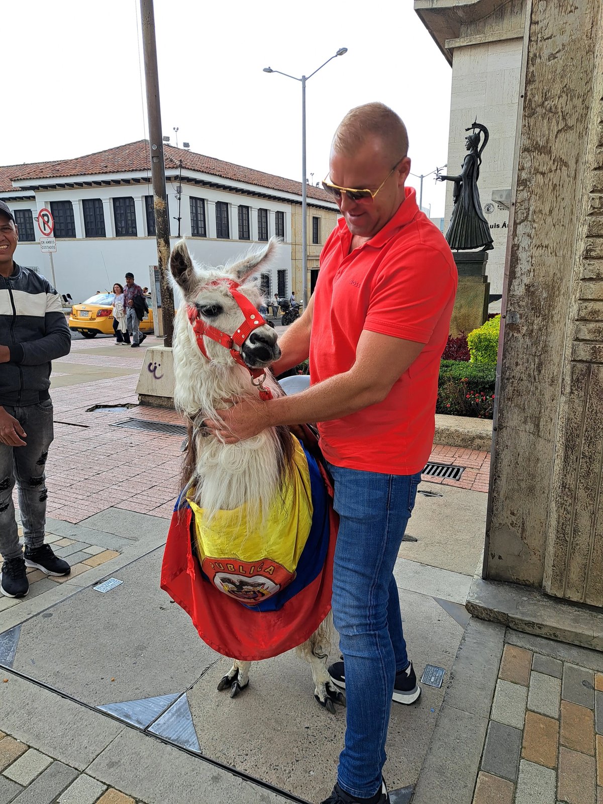 Man in red shirt holding a llama dressed in a colorful cape in a street setting.