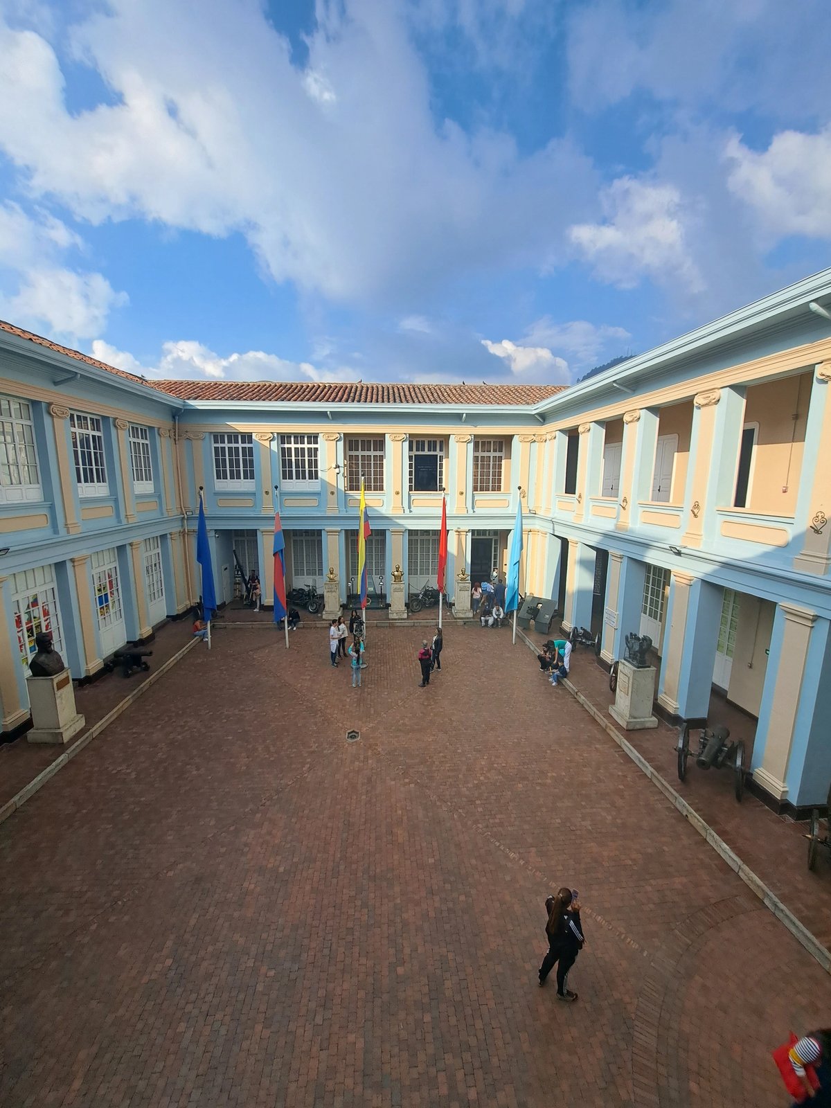 Historic courtyard with flags, busts, and visitors under a blue sky.