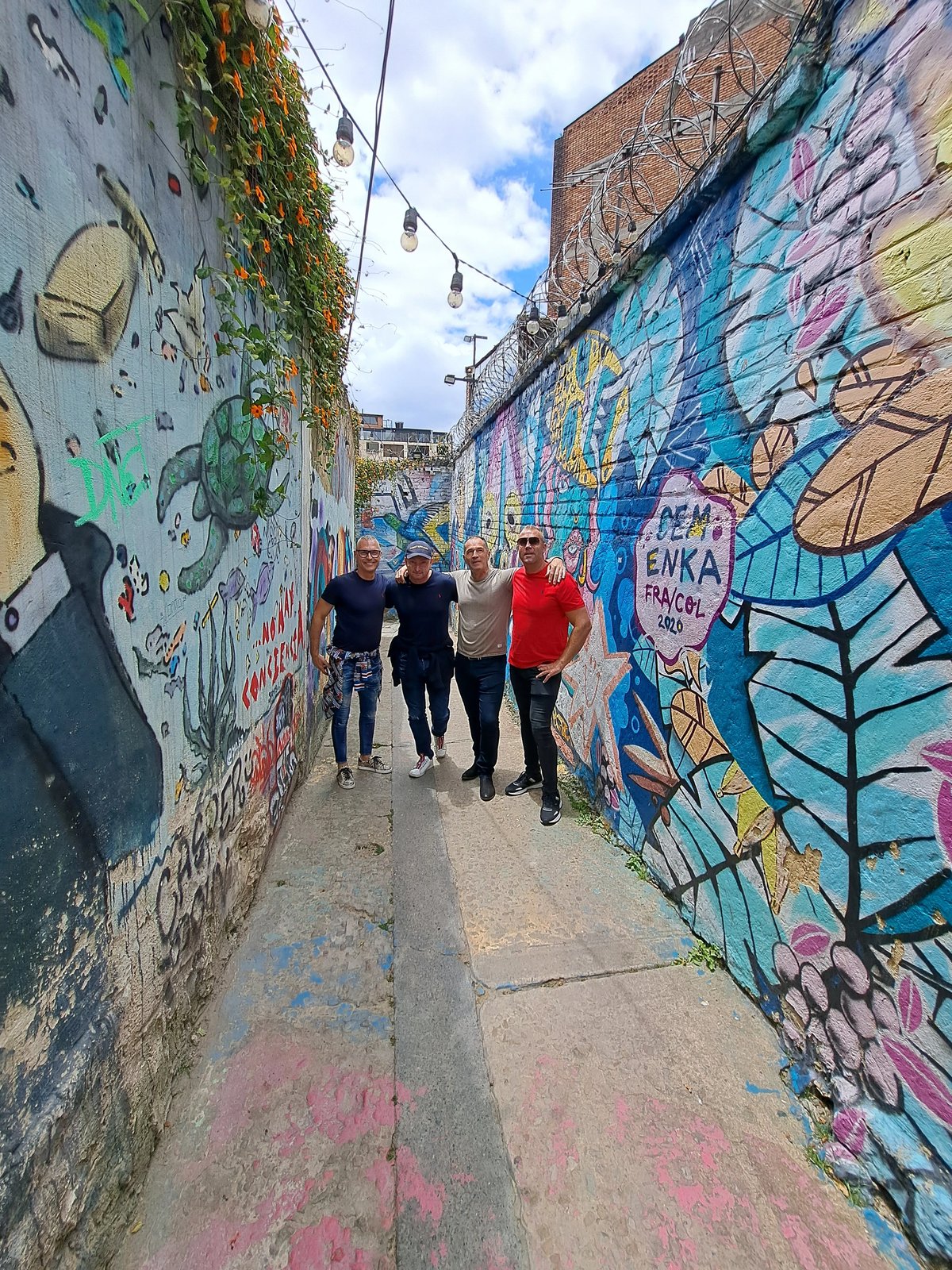 Four men posing in a colorful graffiti-filled alley under a partly cloudy sky.