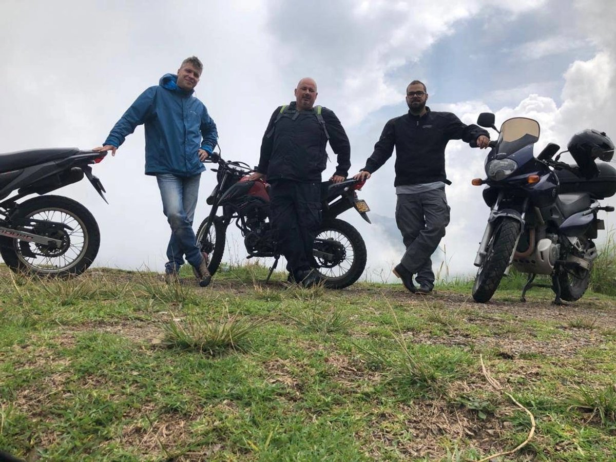 Three men posing with their motorcycles on a grassy hill under a cloudy sky.