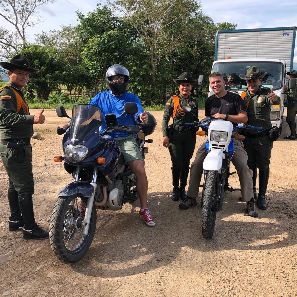 Two men on motorcycles with four police officers giving thumbs up in a rural setting.