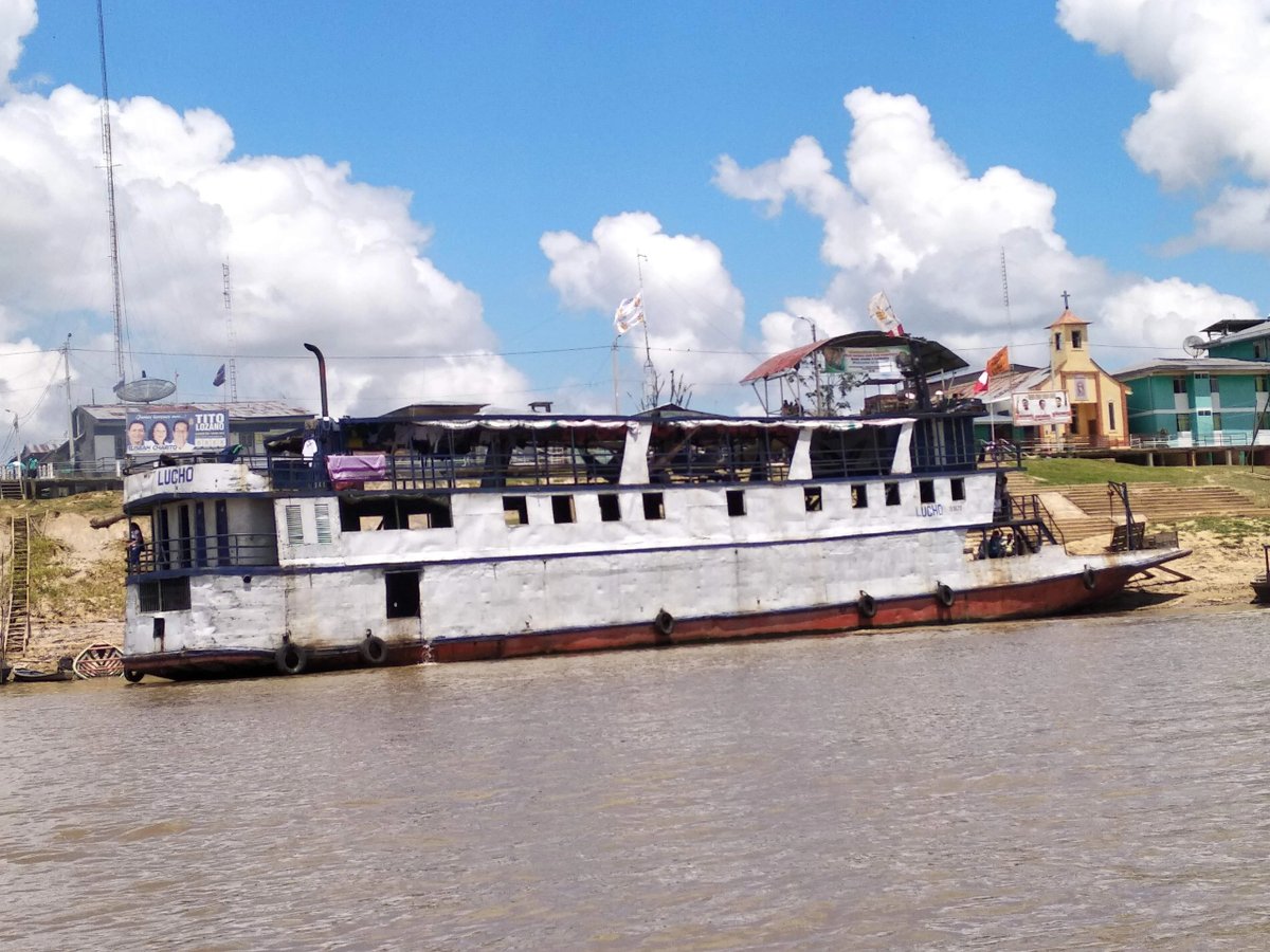 A large white boat named Lucho docked near a riverside village with buildings and a church.
