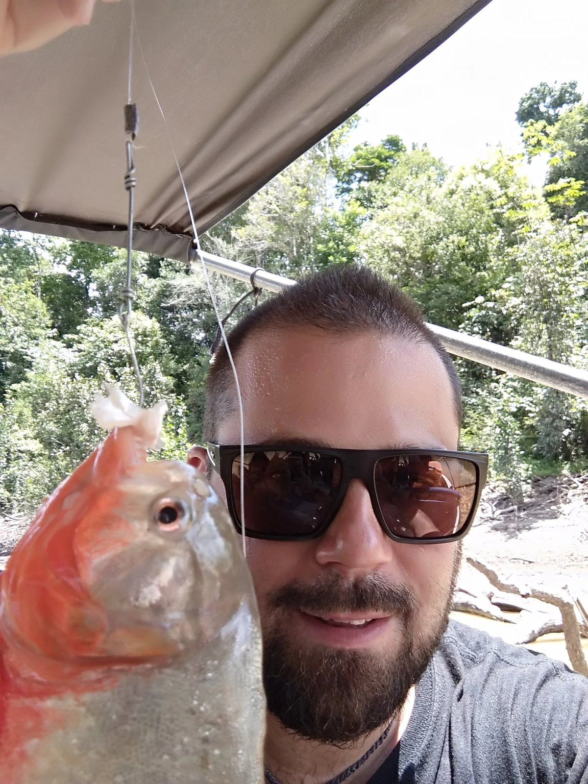 Man wearing sunglasses holding a freshly caught fish on a boat under an awning.