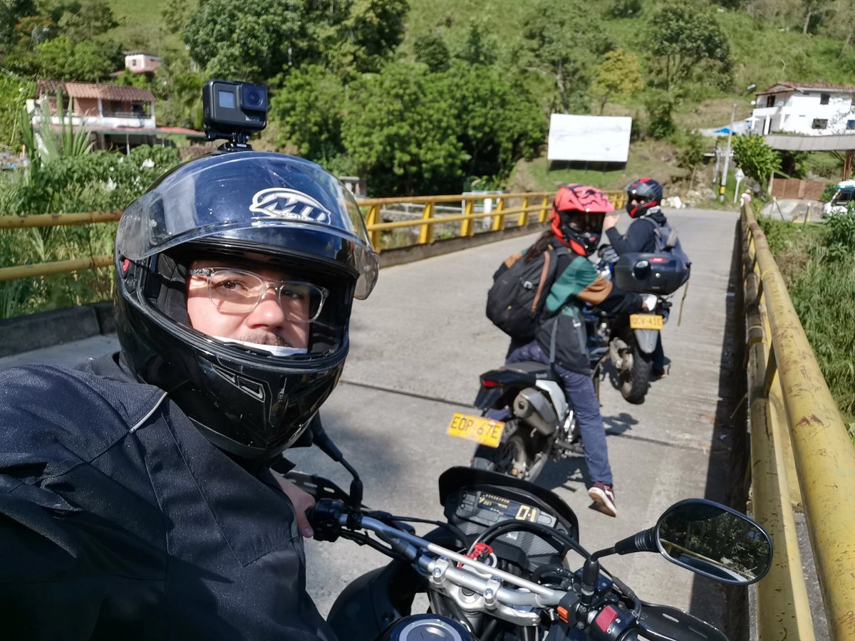 Motorcyclists on a sunny rural bridge, wearing helmets and gear.
