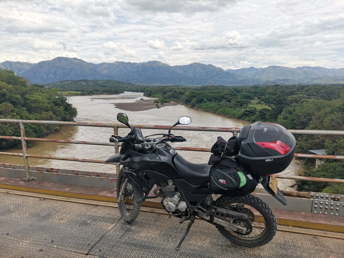 Motorcycle parked on a bridge overlooking a river and distant mountains.