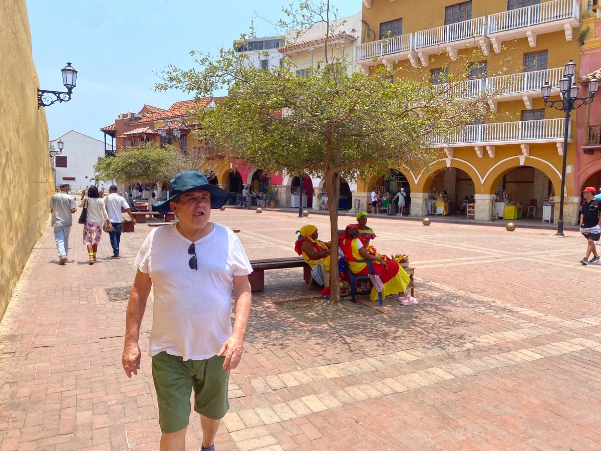 People walking and sitting in a sunny plaza with colorful buildings.
