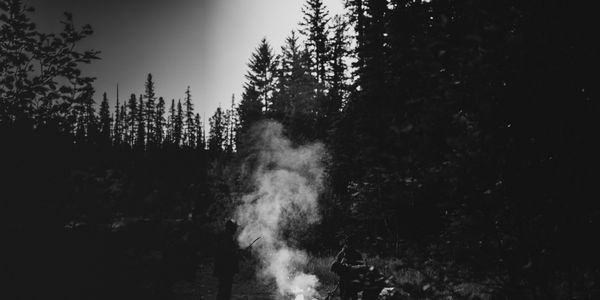 Black and white photo of two people around a campfire in a forest.