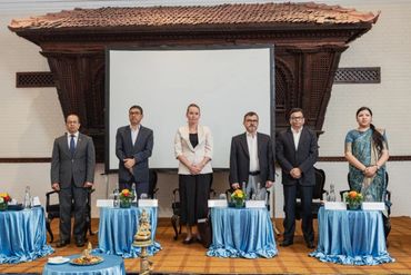 Six professionals standing behind tables with blue cloths in a formal setting.