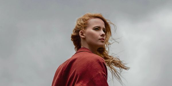 Woman with red hair and red cloak against cloudy sky.