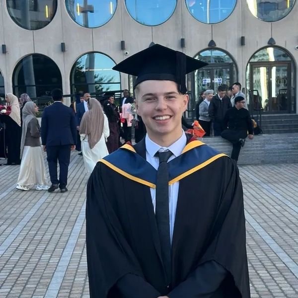A smiling graduate in cap and gown standing outdoors in front of a building.