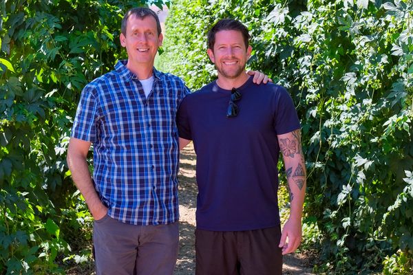 Two men smiling outdoors with greenery in the background.