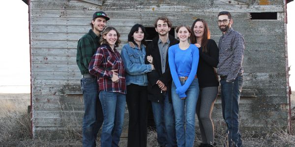 A group of seven people posing outdoors in front of a weathered wooden structure.