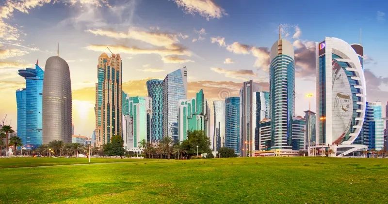Skyline of modern skyscrapers with a green park in the foreground at sunset.