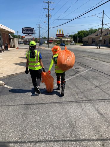 Two women walk on the road with trash bags.