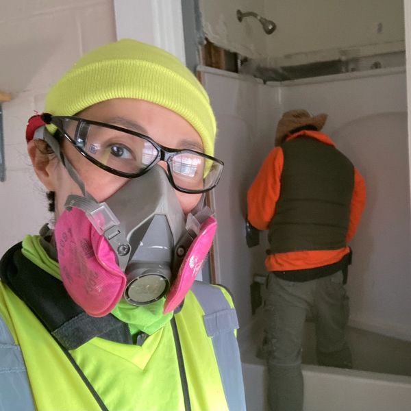A woman faces the camera with her respirator mask while a man works on removing an old bathtub.