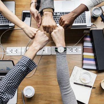 Team of five people fist-bumping over a wooden desk with laptops and notebooks.