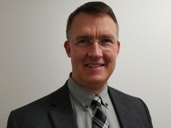 Man in glasses wearing a gray suit and plaid tie smiles at the camera.