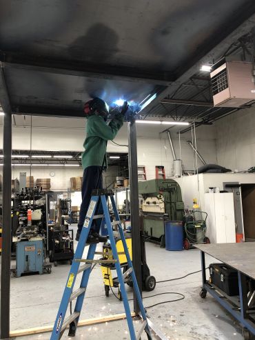 Worker welding metal while standing on a ladder in workshop.