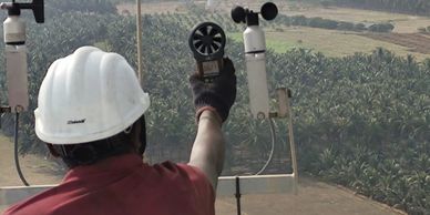 Technician in a hard hat measuring wind speed near wind turbines with an anemometer.