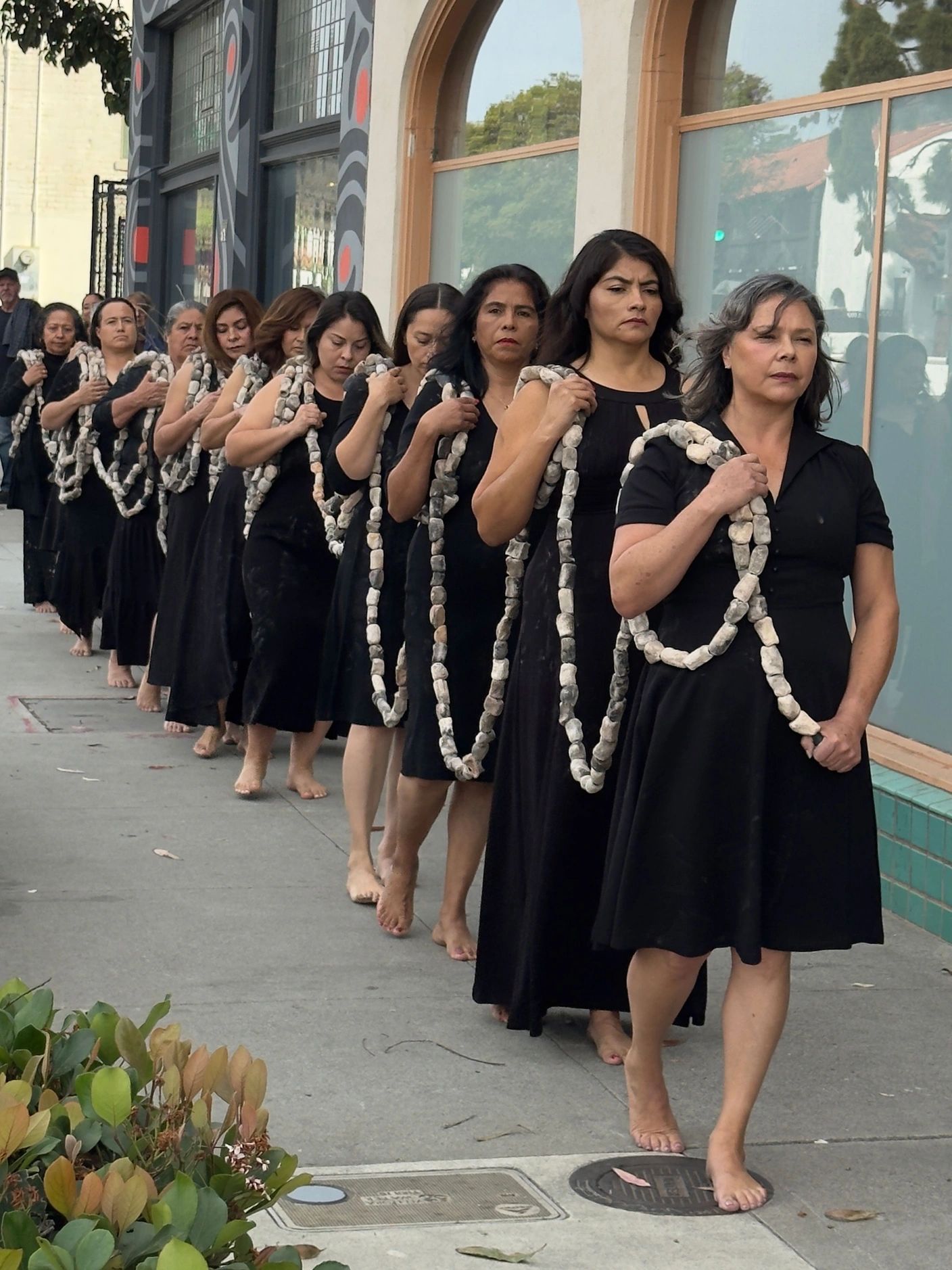 A line of barefoot women in black dresses holding stone necklaces outside an art center.