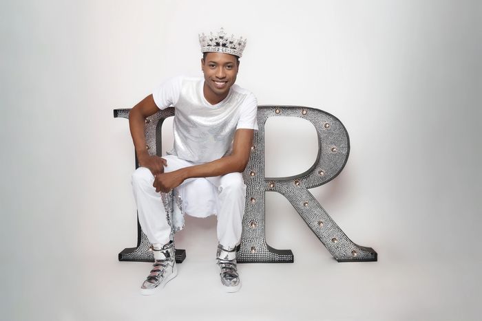 Howard Reese smiling in a crown, dressed in white, seated on an “HR” prop against a white background