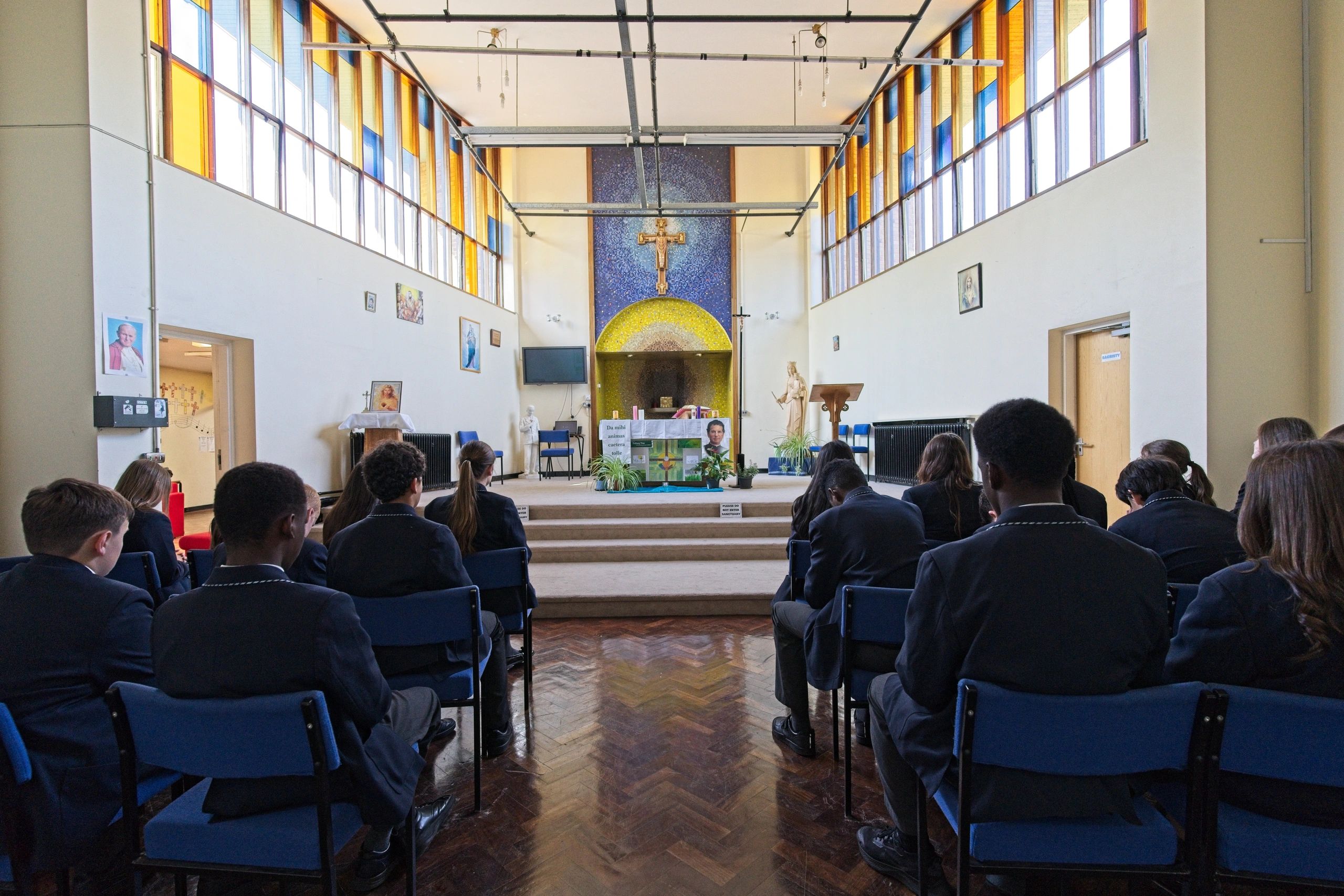 Students seated in a chapel with stained glass windows and religious symbols.