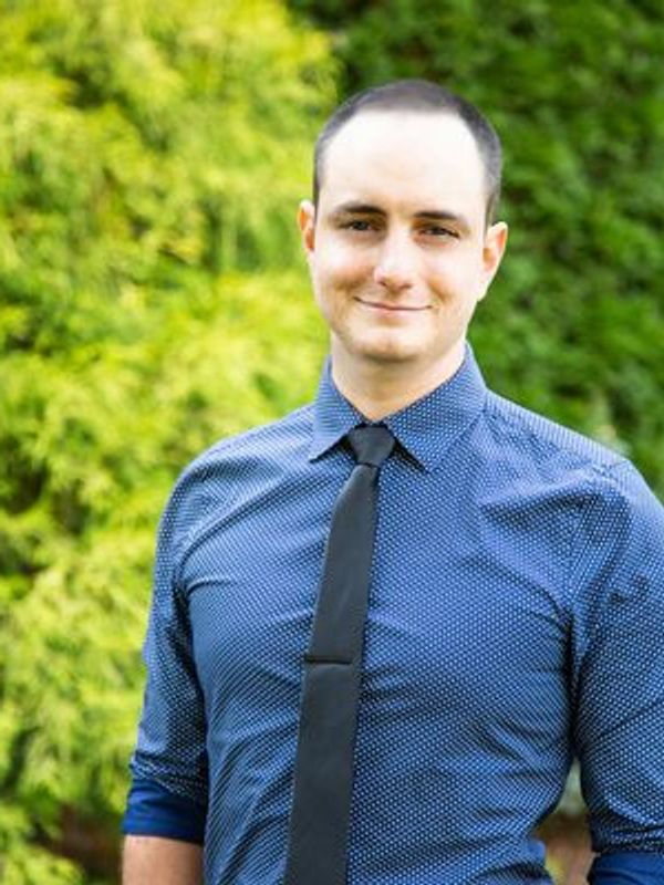 Man in a blue patterned shirt and black tie smiling outdoors.